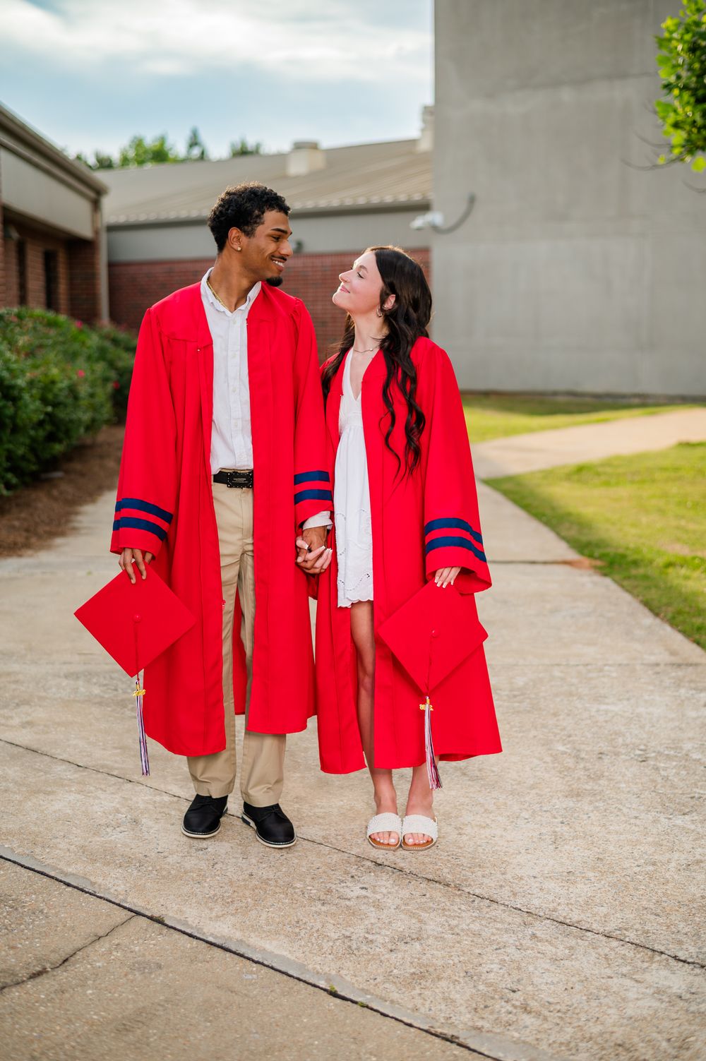 Two graduates in red caps and gowns hold hands and smile at each other outdoors on a sunny day.