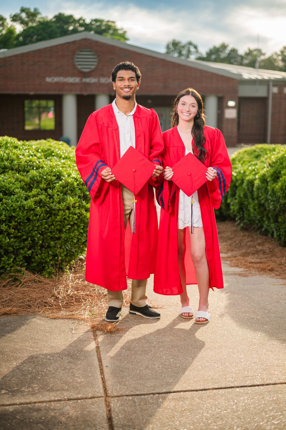 Two high school graduates in red caps and gowns pose proudly outside Northside High School on graduation day.