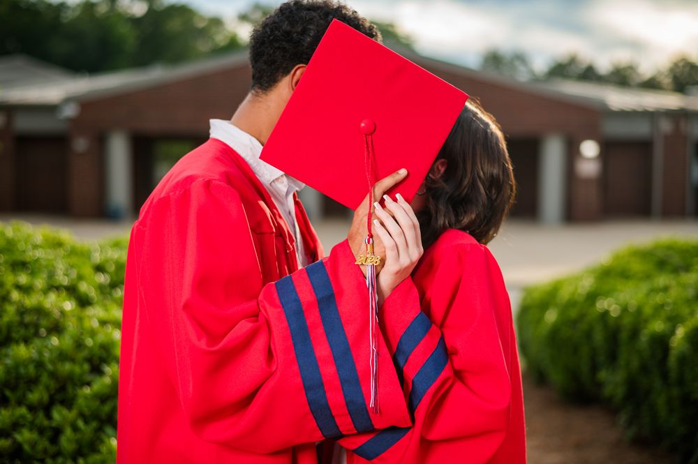 Two graduates in red caps and gowns share a kiss, hiding their faces behind a graduation cap outdoors.