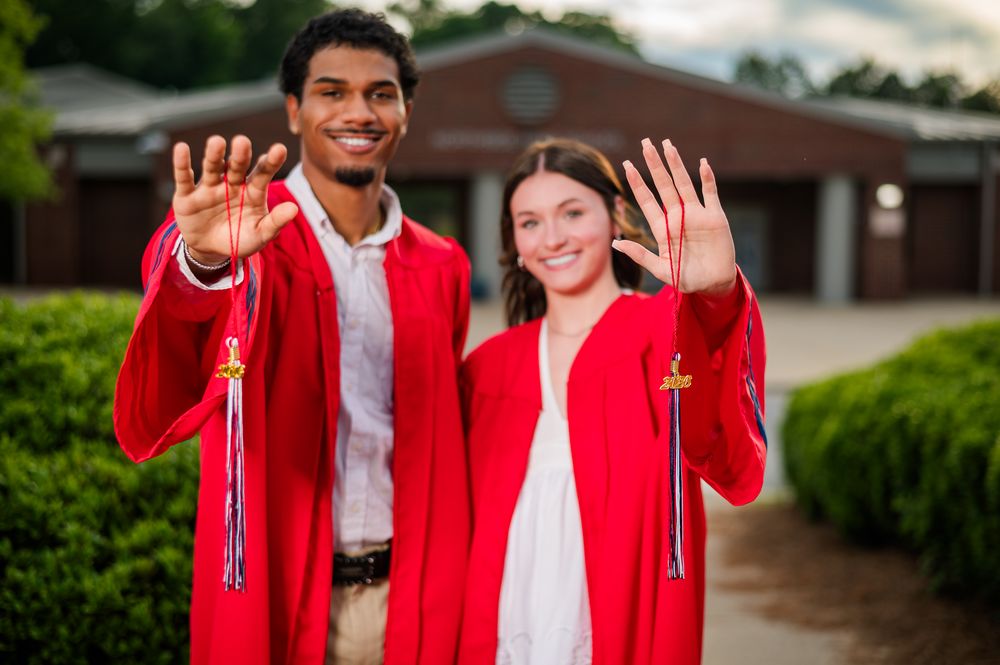 Two graduates in red gowns and tassels smile while showing their hands to the camera outdoors.