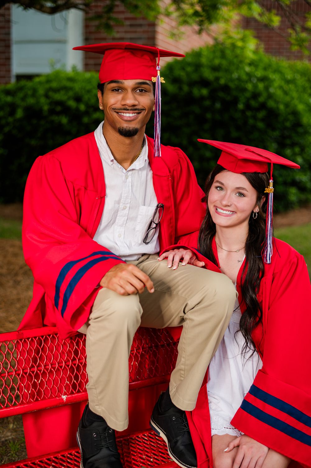 Two graduates in red caps and gowns smiling on a red bench outdoors.