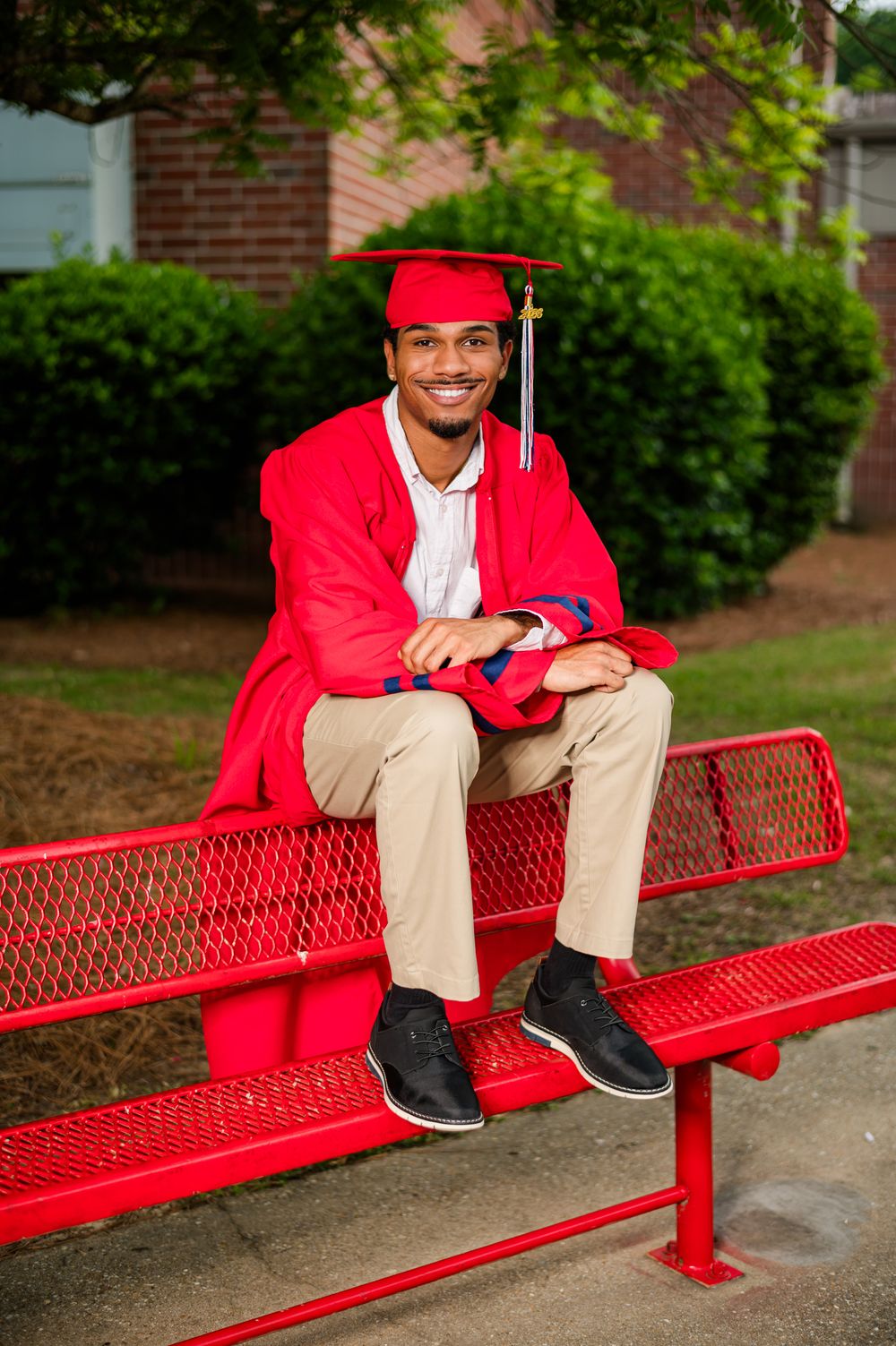 Smiling graduate in red cap and gown sitting on a red bench outdoors, celebrating high school or college graduation.