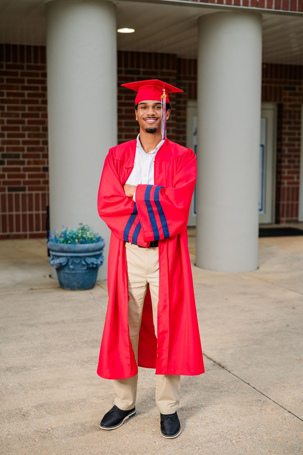 Young man in red graduation cap and gown stands proudly with arms crossed outside a brick building.