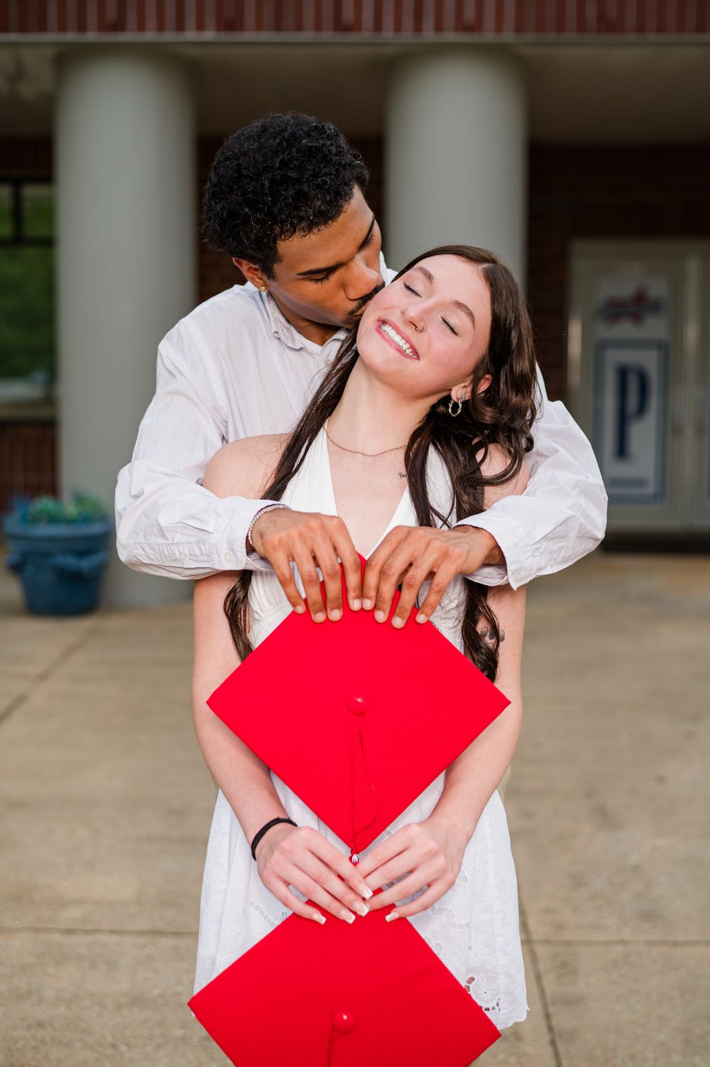 A couple celebrates graduation, sharing a kiss while holding a red graduation cap outside.