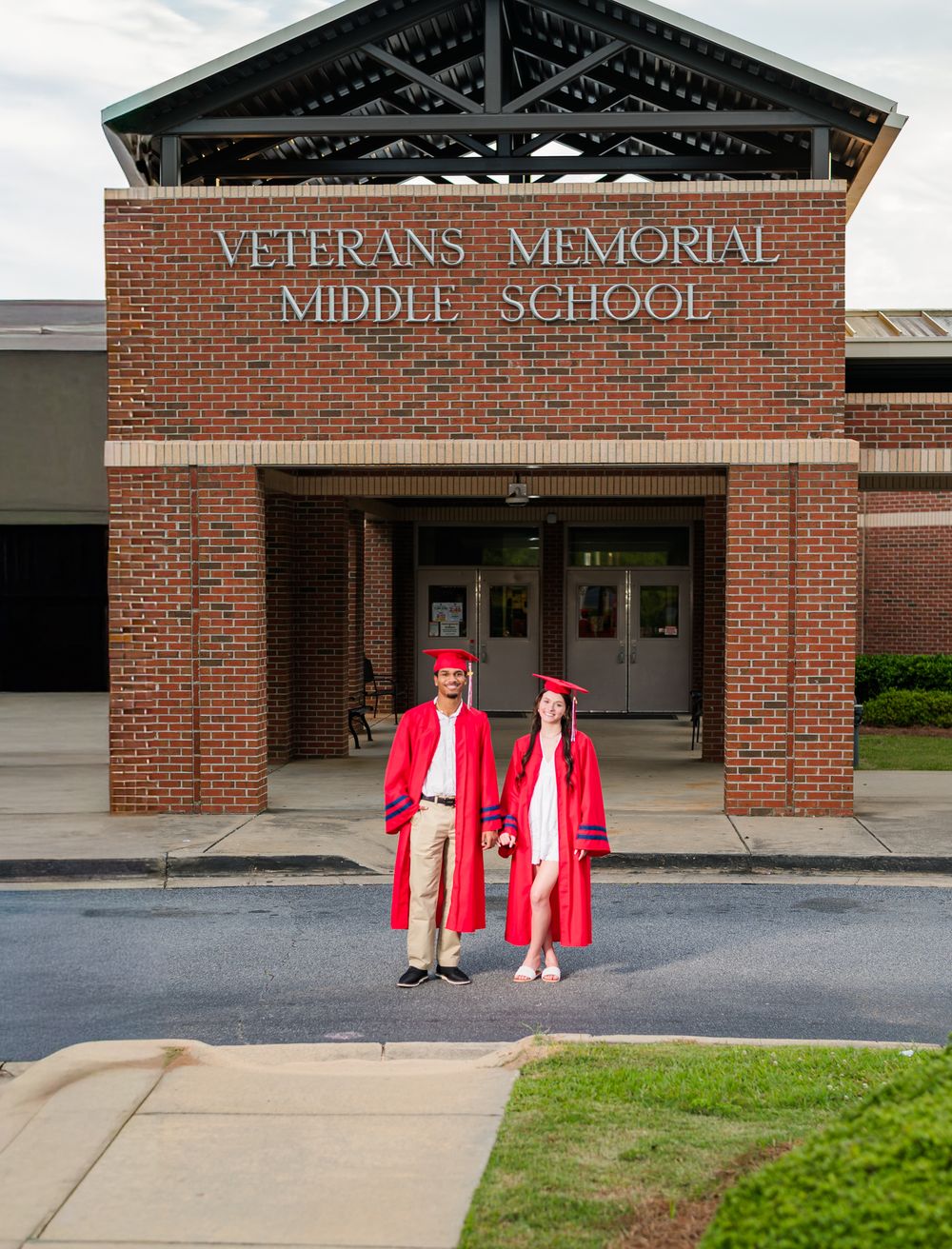 Two graduates in red cap and gown pose proudly in front of Veterans Memorial Middle School on graduation day.
