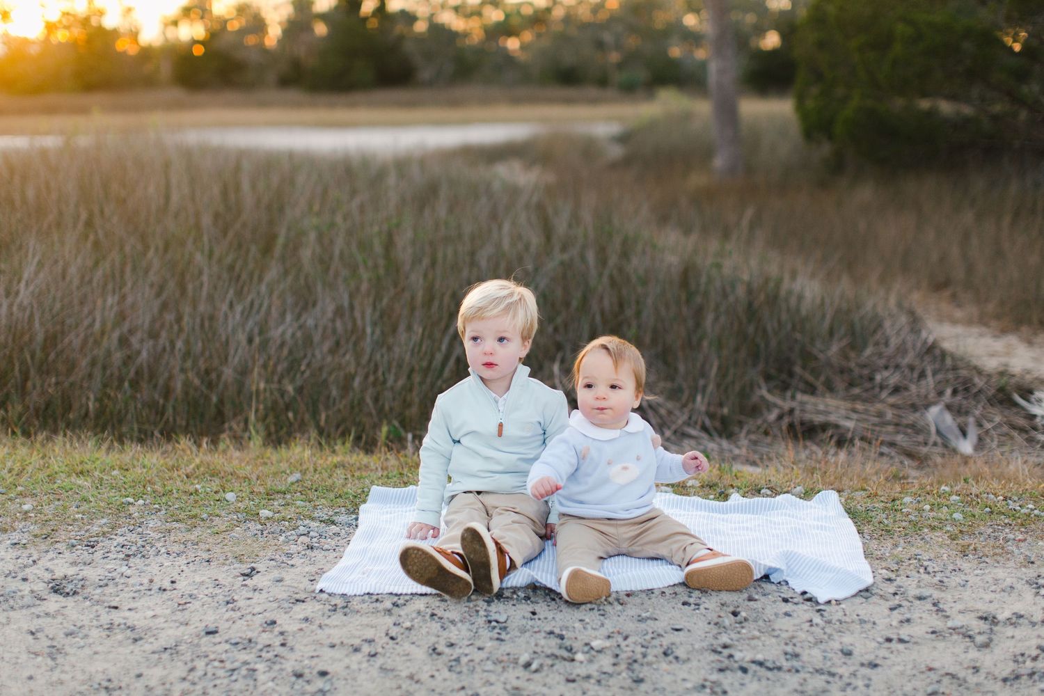 Two young siblings from Hilton Head, SC sit together for family photography on a white blanket at a sandy beach during golden hour sunset.