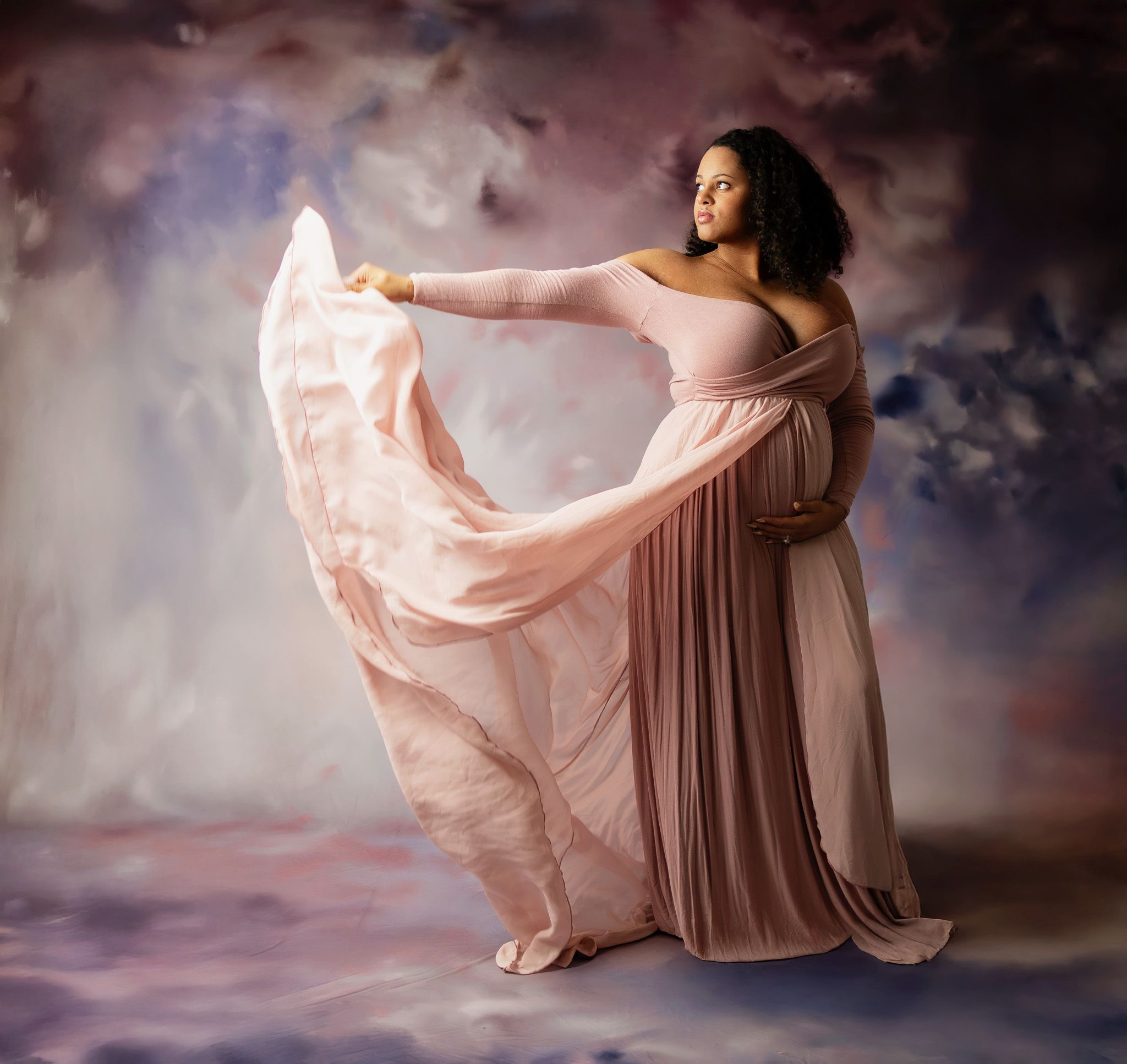 Pregnant woman in flowing pink maternity gown poses gracefully against a dreamy purple and blue backdrop.