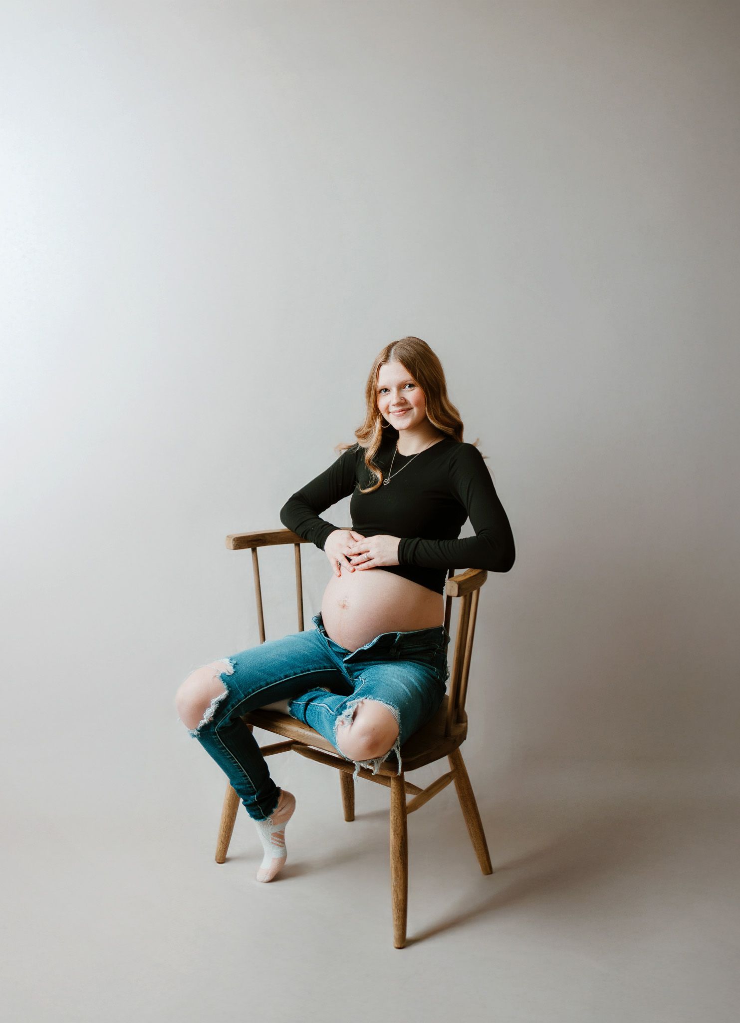 Smiling pregnant woman in black top and ripped jeans sitting on wooden chair, cradling baby bump in studio maternity photo.