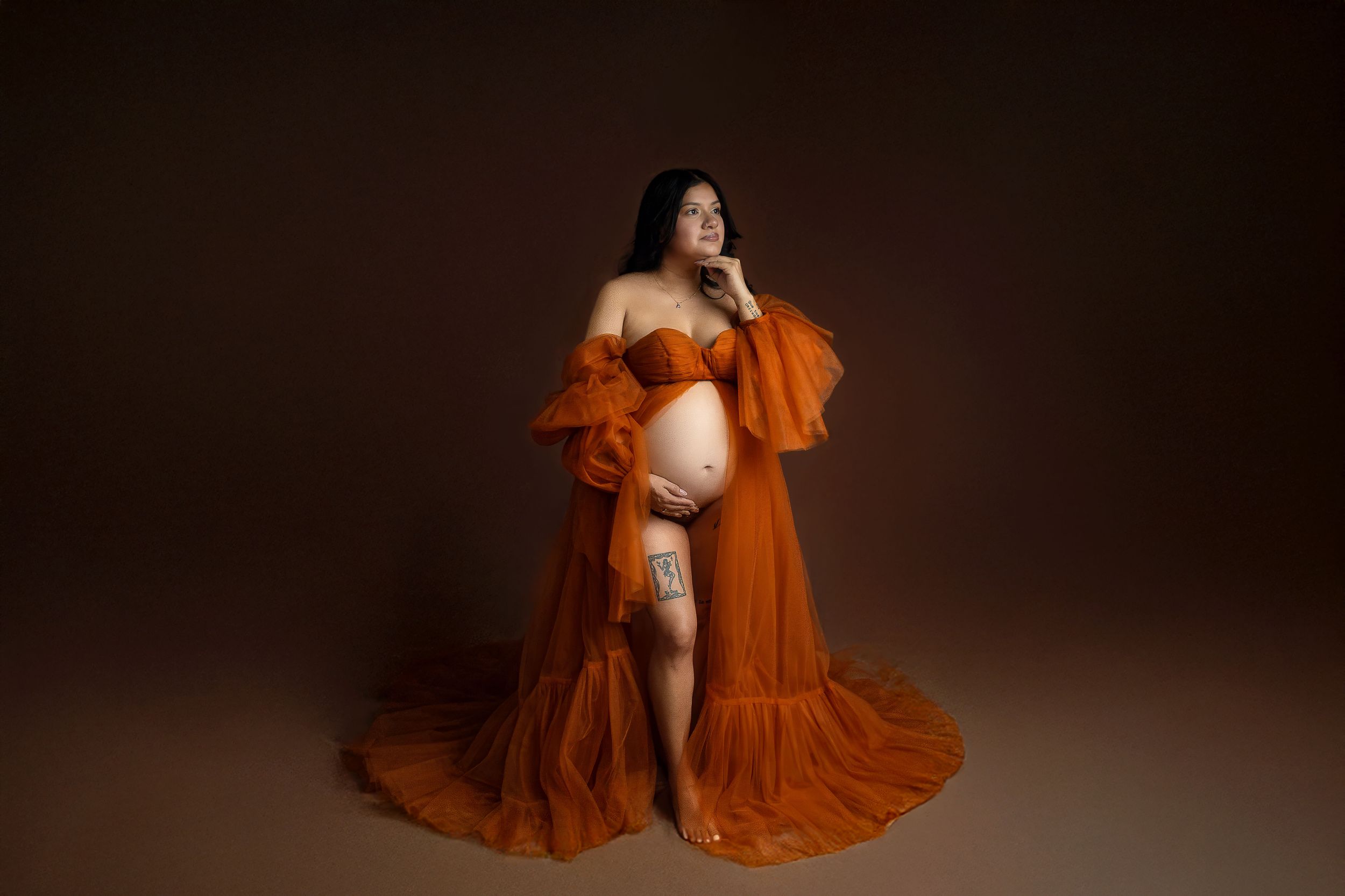 Pregnant woman in elegant rust orange tulle maternity gown posing gracefully in a professional studio photo session.