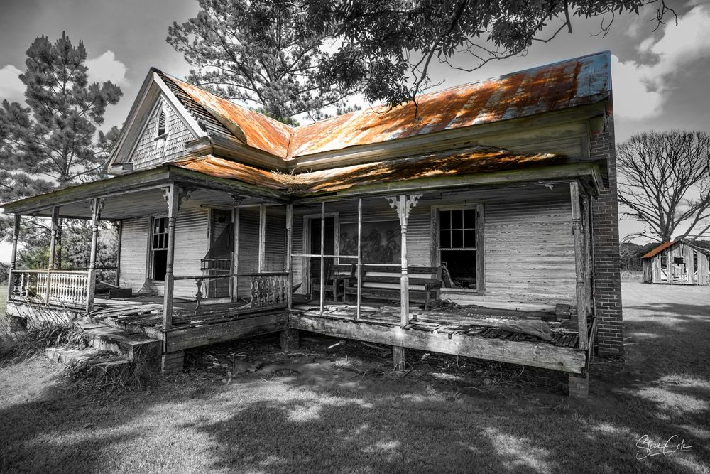 Black and white photo with selective color highlighting the rusted tin roof of an old, abandoned farmhouse, photo by Steve Cole Photography.