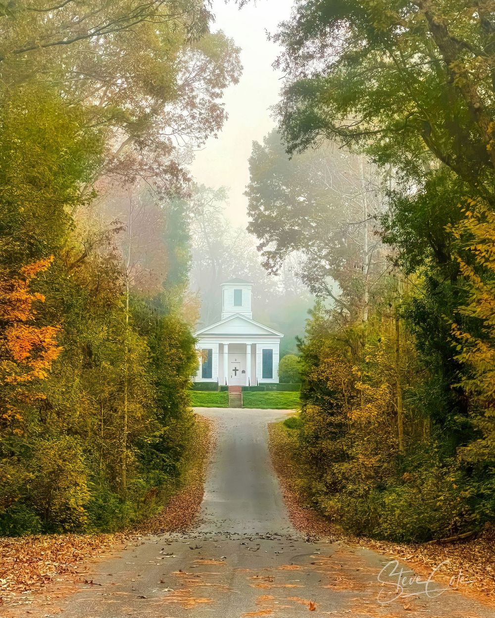 Country road lined with autumn trees leading to a historic white church shrouded in light fog, photo by Steve Cole Photography.