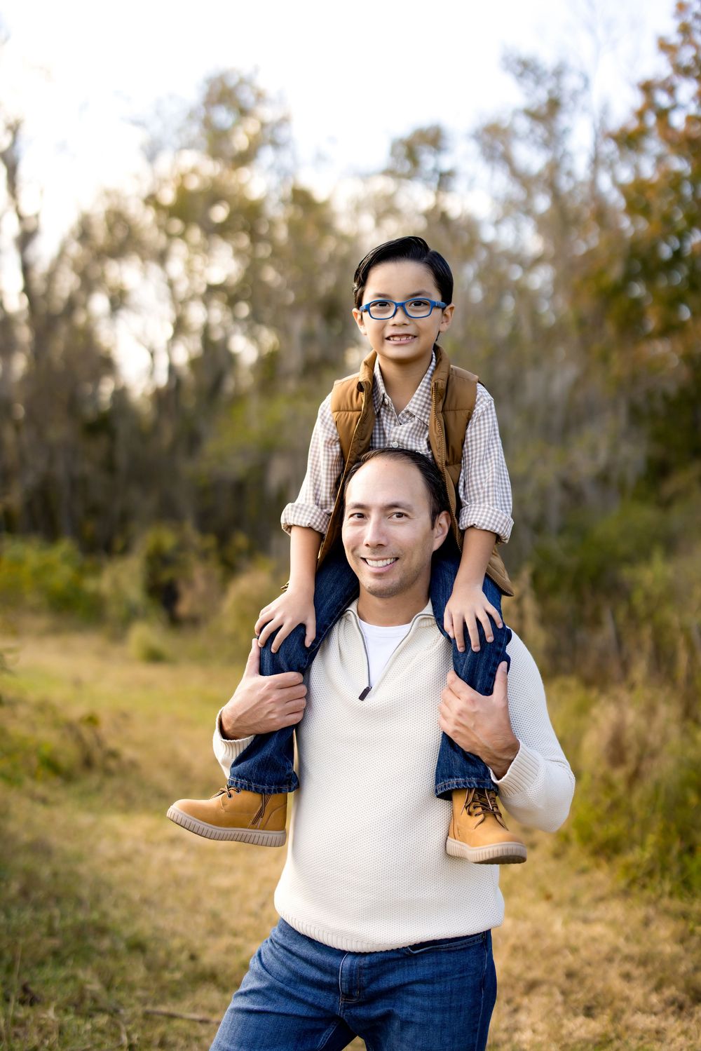A smiling man carries a young boy with glasses on his shoulders in an autumn outdoor setting.