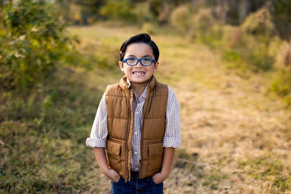 Smiling young boy wearing glasses and a brown vest standing in a sunny outdoor field.