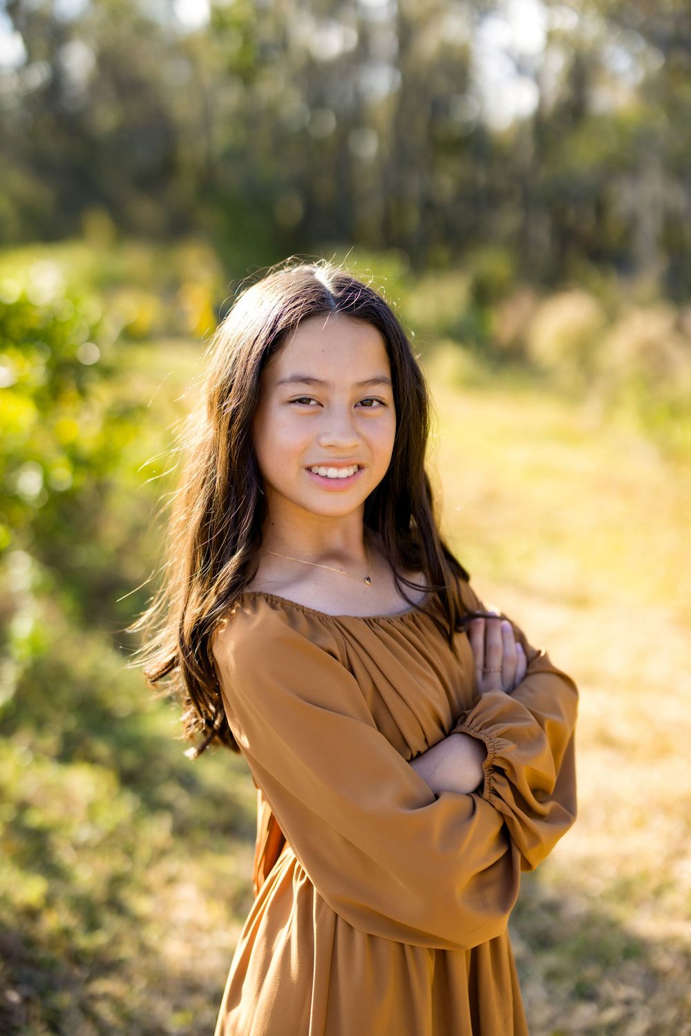 A smiling young girl with long dark hair wearing a golden off-shoulder dress stands outdoors with arms crossed.