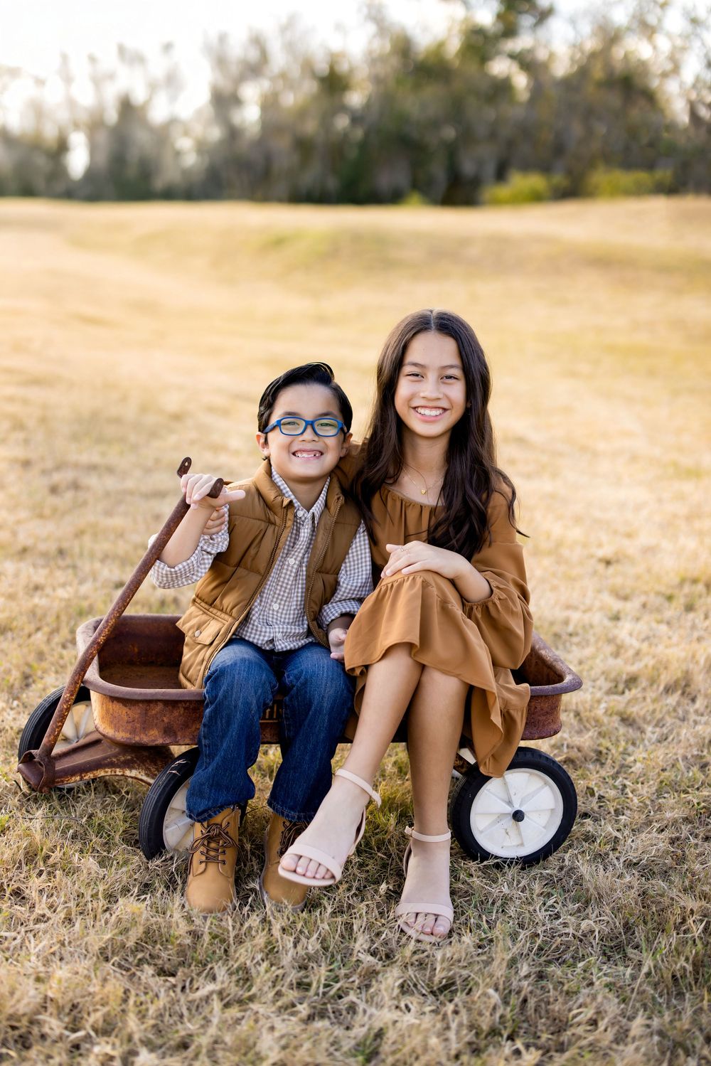 Two smiling children sit together in a vintage red wagon in a sunny field, the girl in a brown dress and boy in glasses.