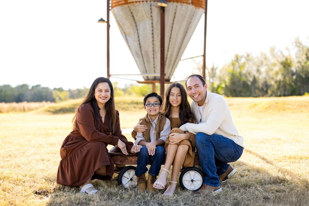 Family of four posing outdoors near a vintage hot air balloon prop on a wagon in a sunny field.