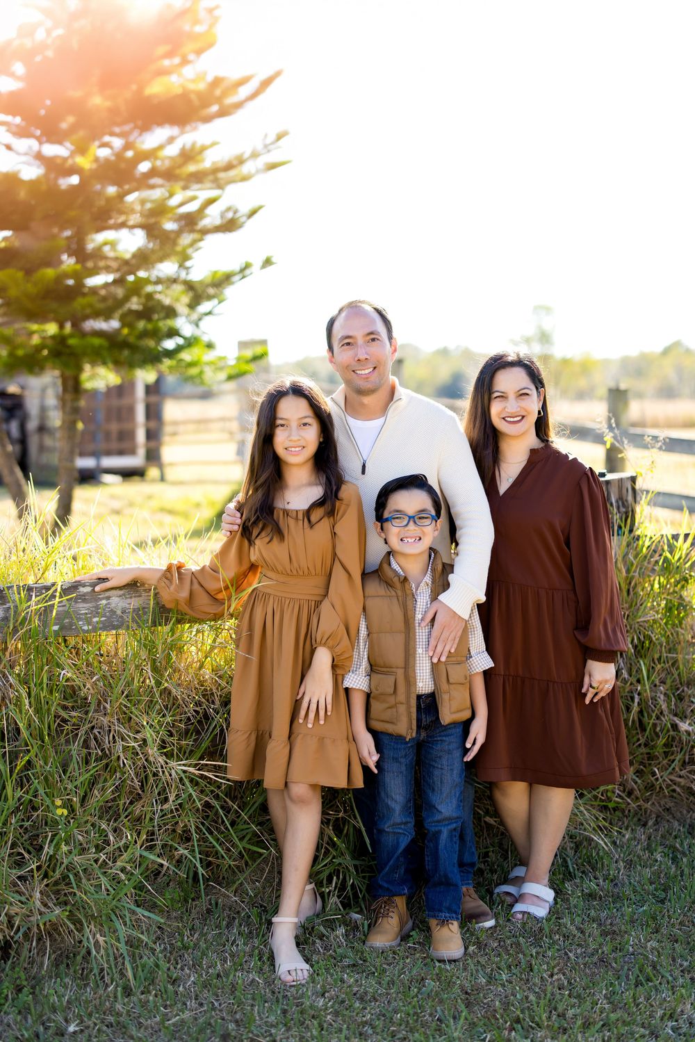 Family of four posing outdoors near a wooden fence, wearing warm earth tones, smiling in golden afternoon light.