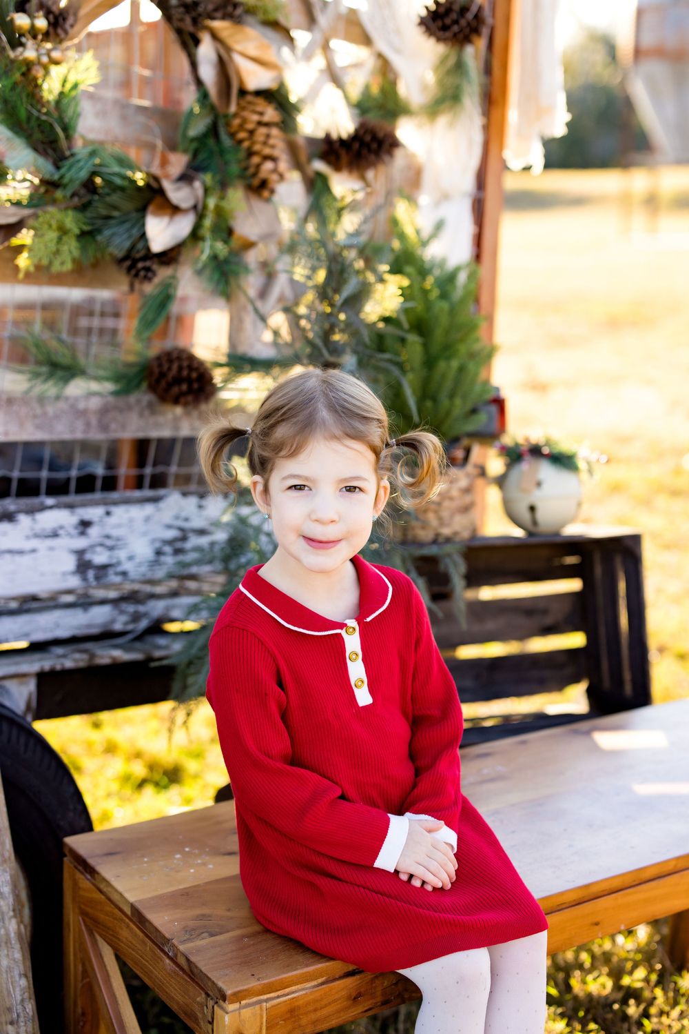 Young girl in red dress sitting on wooden table with Christmas wreath decor backdrop outdoors.
