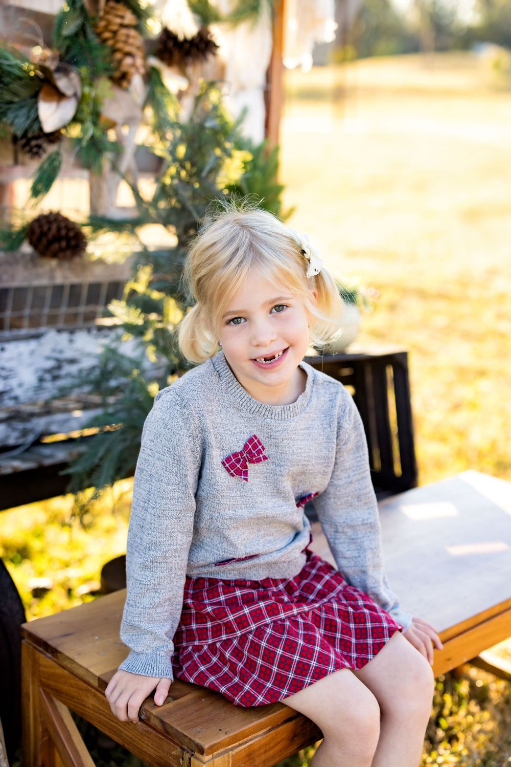 Smiling blonde girl in gray sweater and plaid skirt sits on wooden box near Christmas greenery outdoors.