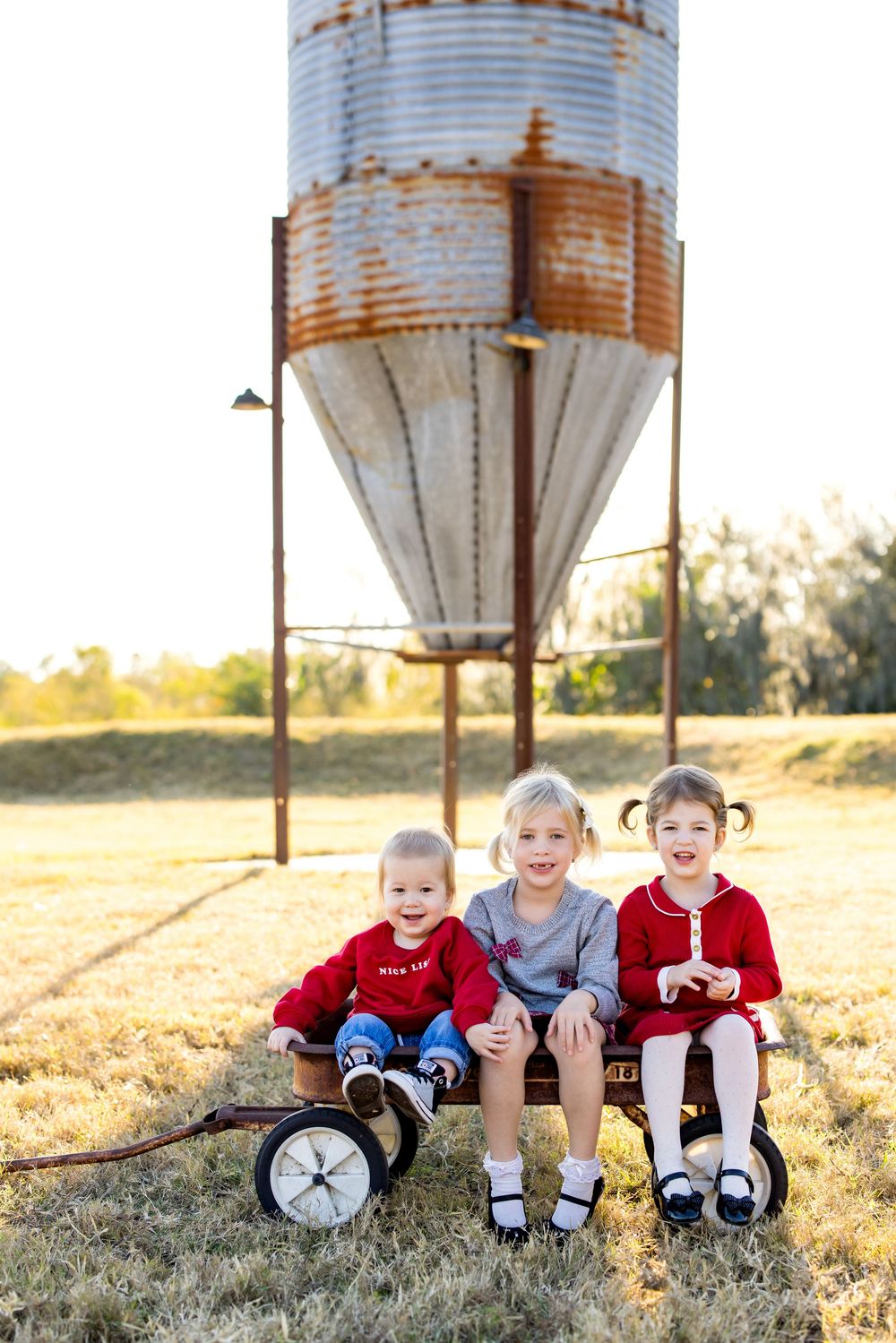 Three young children sit together on a vintage wagon in a rural field with a rusty grain silo in the background.