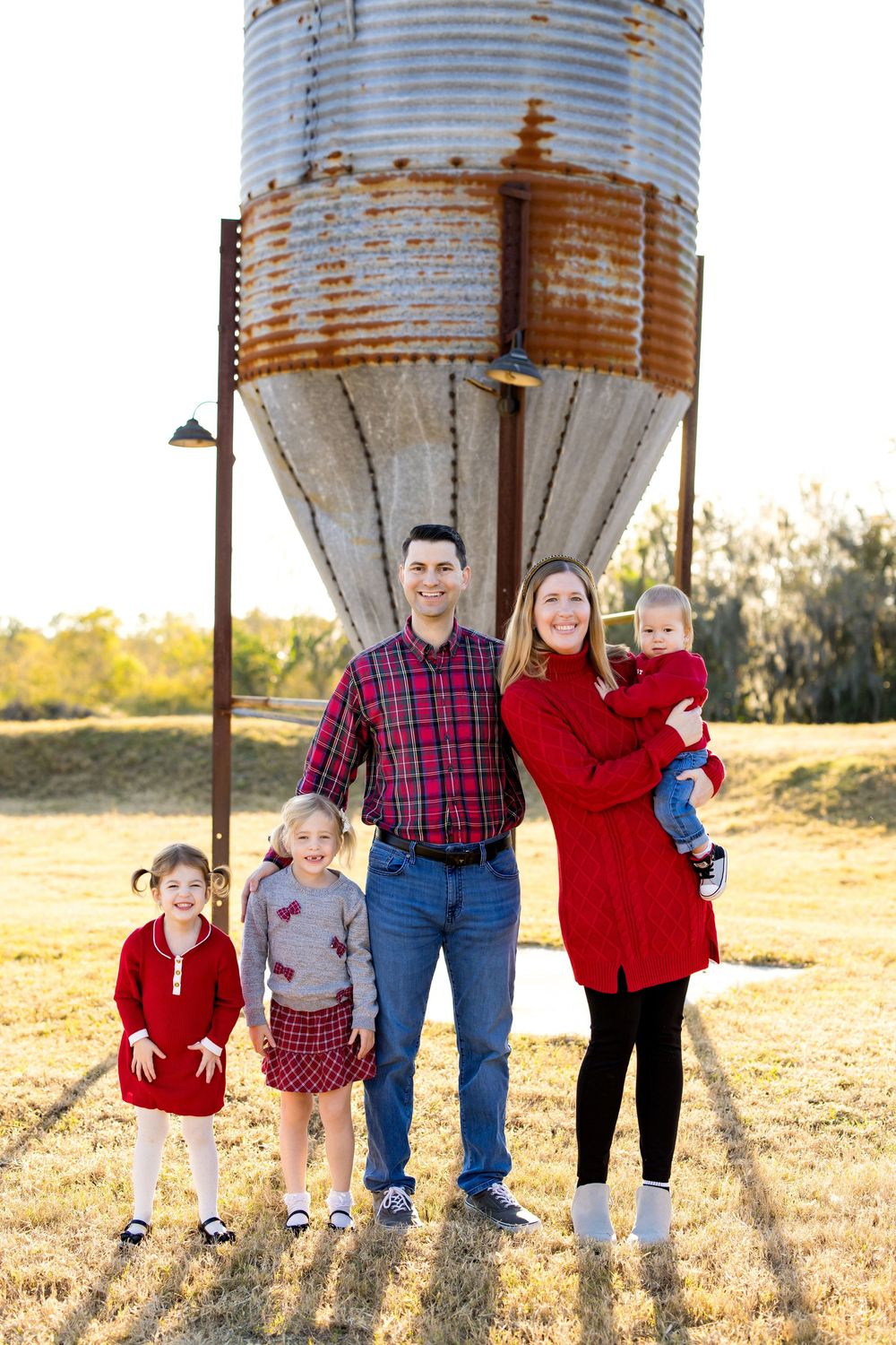 Family of five posing outdoors near a rustic grain silo, wearing coordinated red outfits in a golden field.