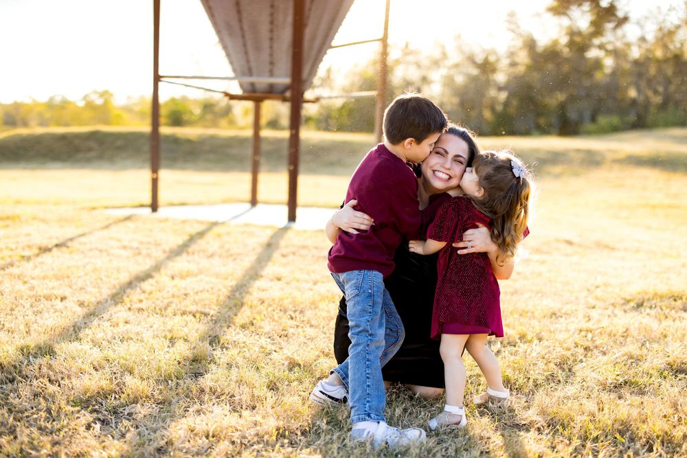 A mother hugs her two children in a sunny field, all wearing matching burgundy outfits, with a structure in the background.