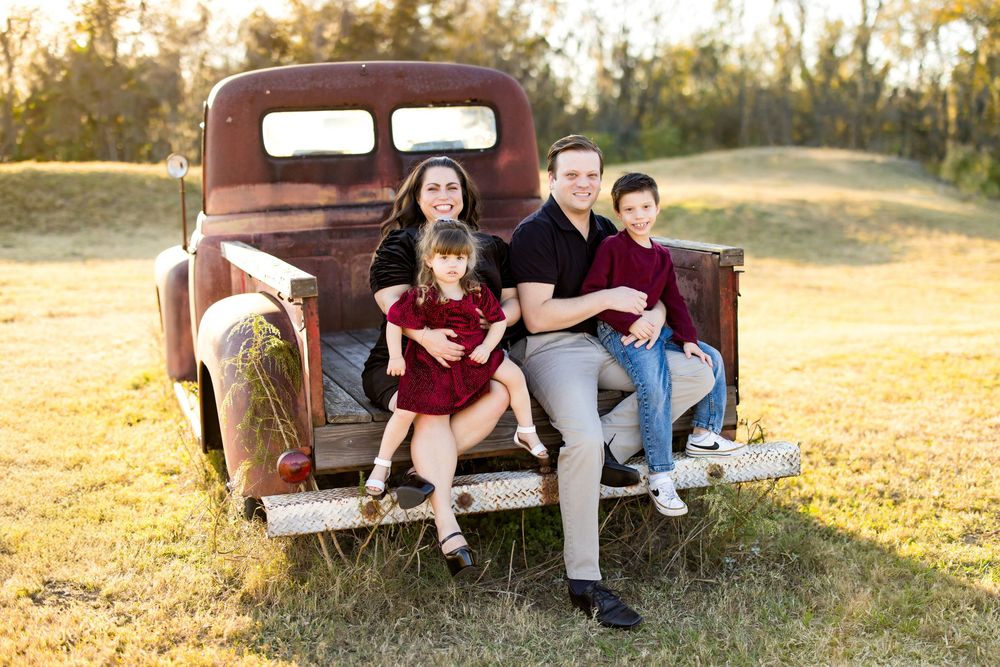 Family of four posing on vintage rusty truck bed in golden hour outdoor setting.