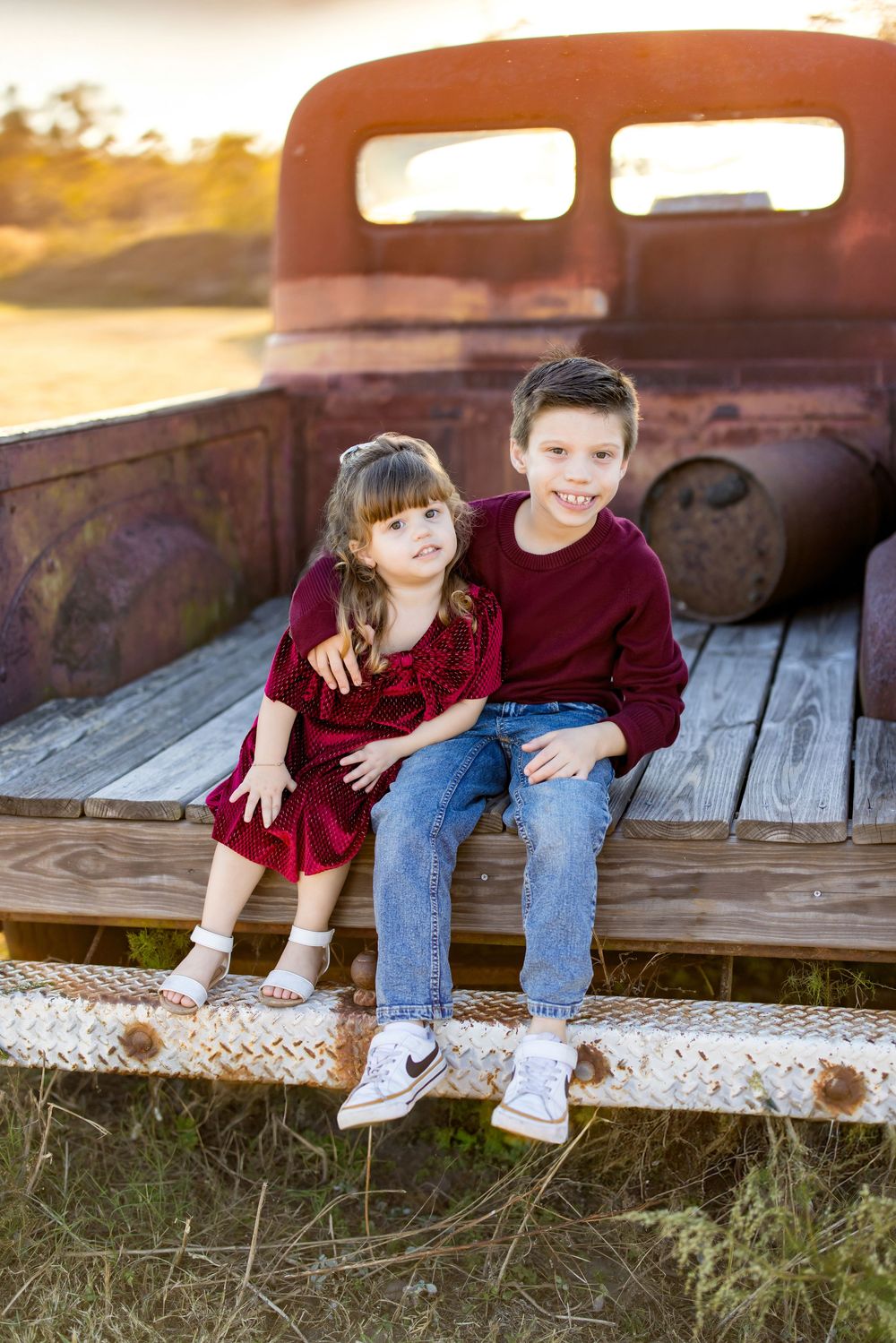 Two children in matching burgundy outfits sit on the tailgate of a rustic vintage truck at golden hour.