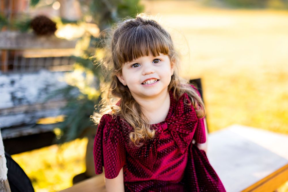 Smiling young girl with curly hair wearing a red velvet dress, sitting outdoors in warm golden light.