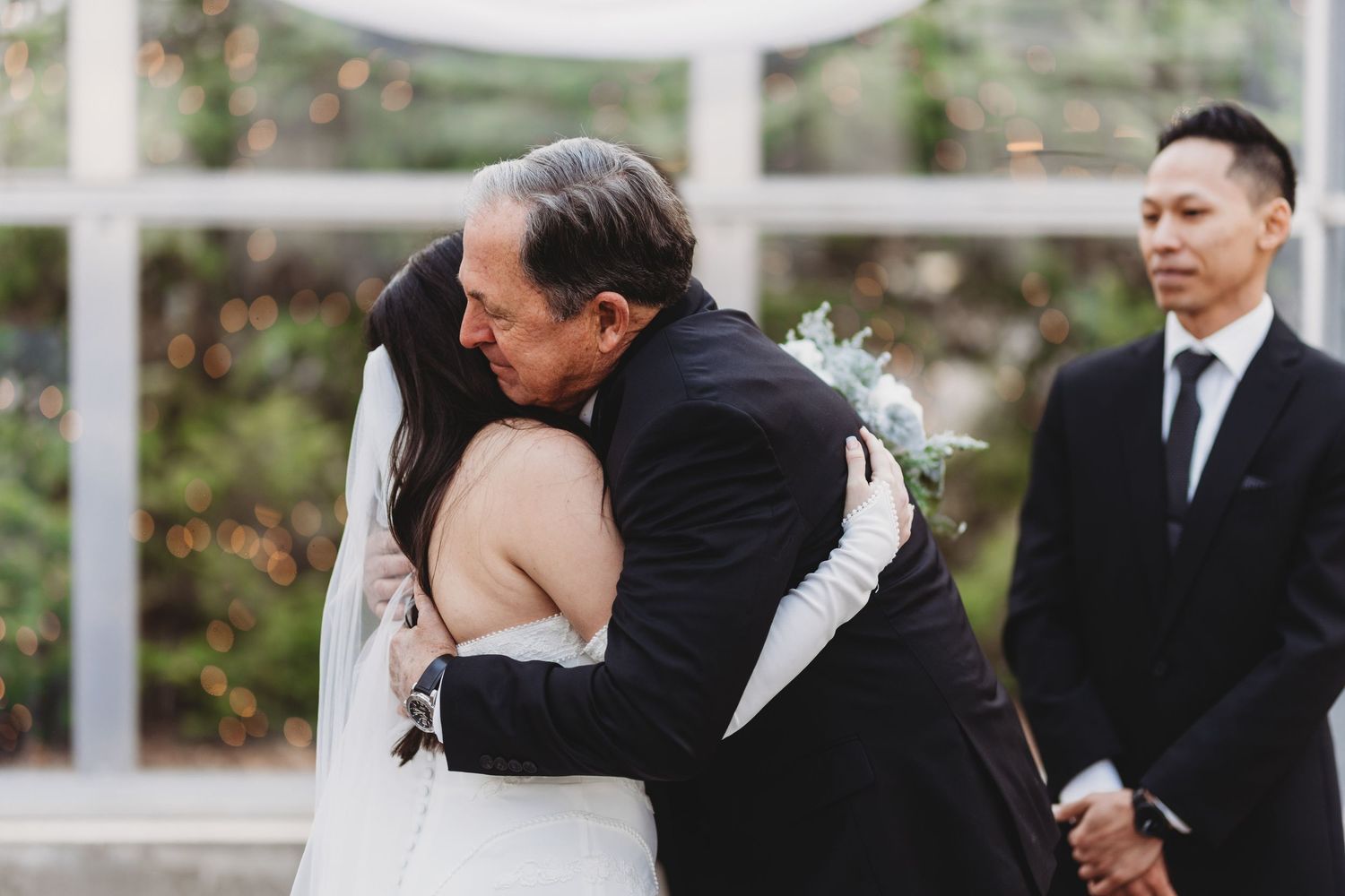 Bride and groom exchange vows in elegant glass chapel surrounded by lush forest trees during intimate wedding ceremony.