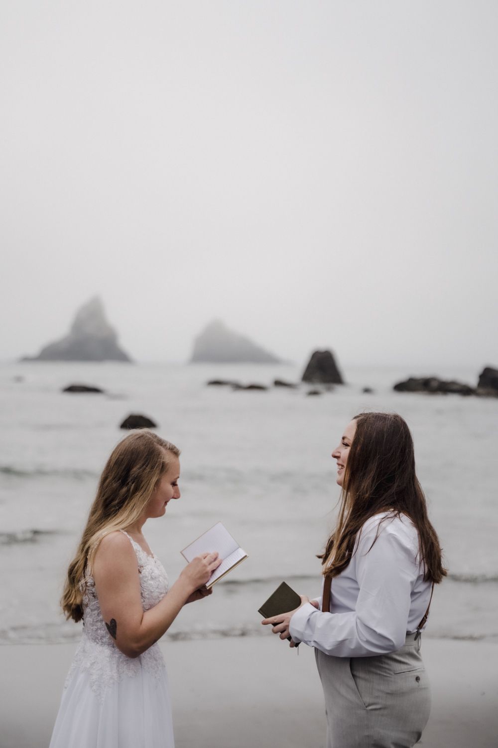 Two brides smile and exchange vows while holding vow books on the beach in front of sea stacks on the Oregon Coast