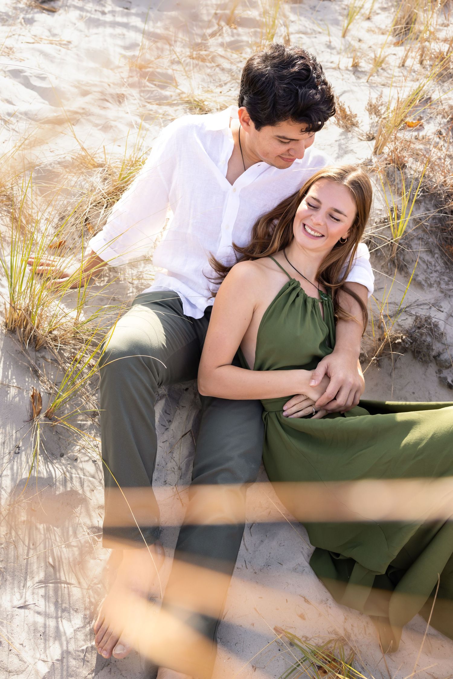 Romantic couple sitting together on sandy beach dunes, woman in green dress laughing joyfully.