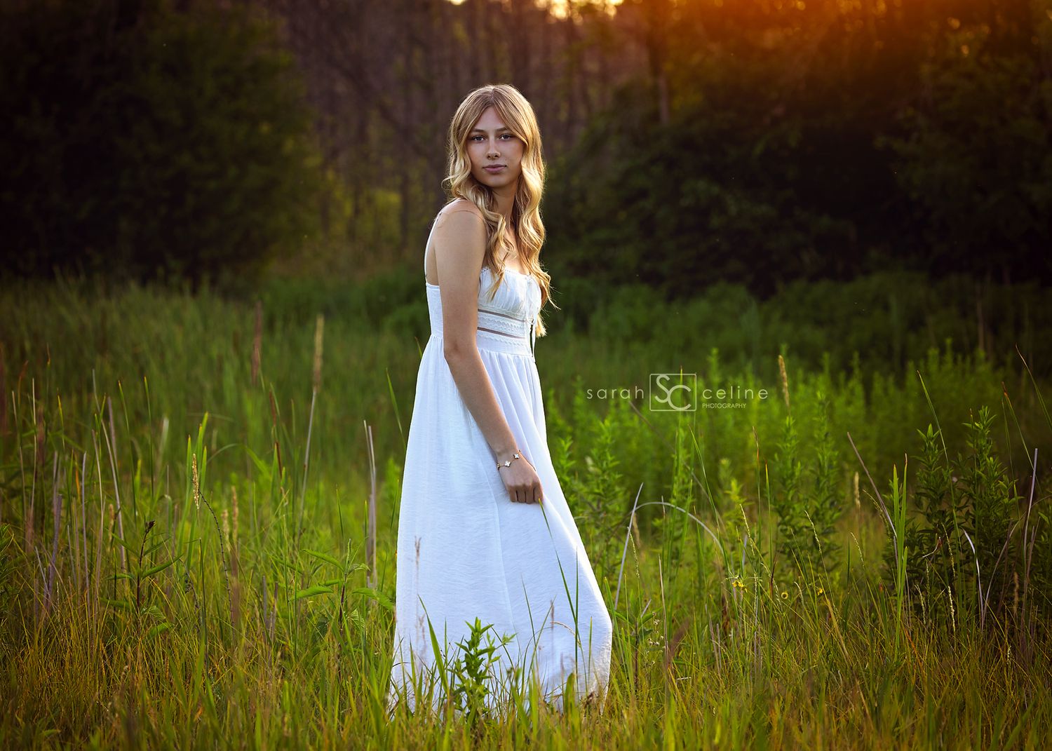 Young blonde woman in white maxi dress standing in lush green field during golden hour sunset.
