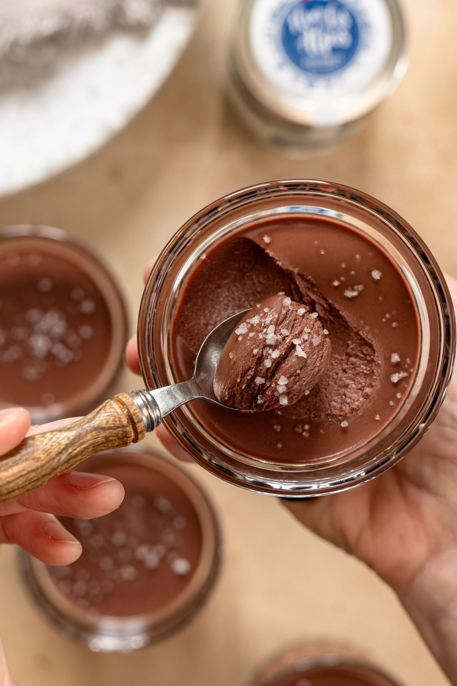 Hand scooping rich chocolate mousse with sea salt flakes from a glass jar, with more jars in background.