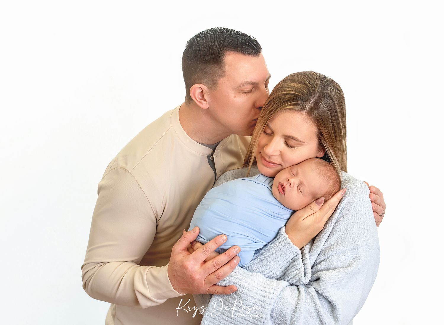 A couple shares a tender moment kissing while cradling their sleeping newborn baby wrapped in a blue swaddle.