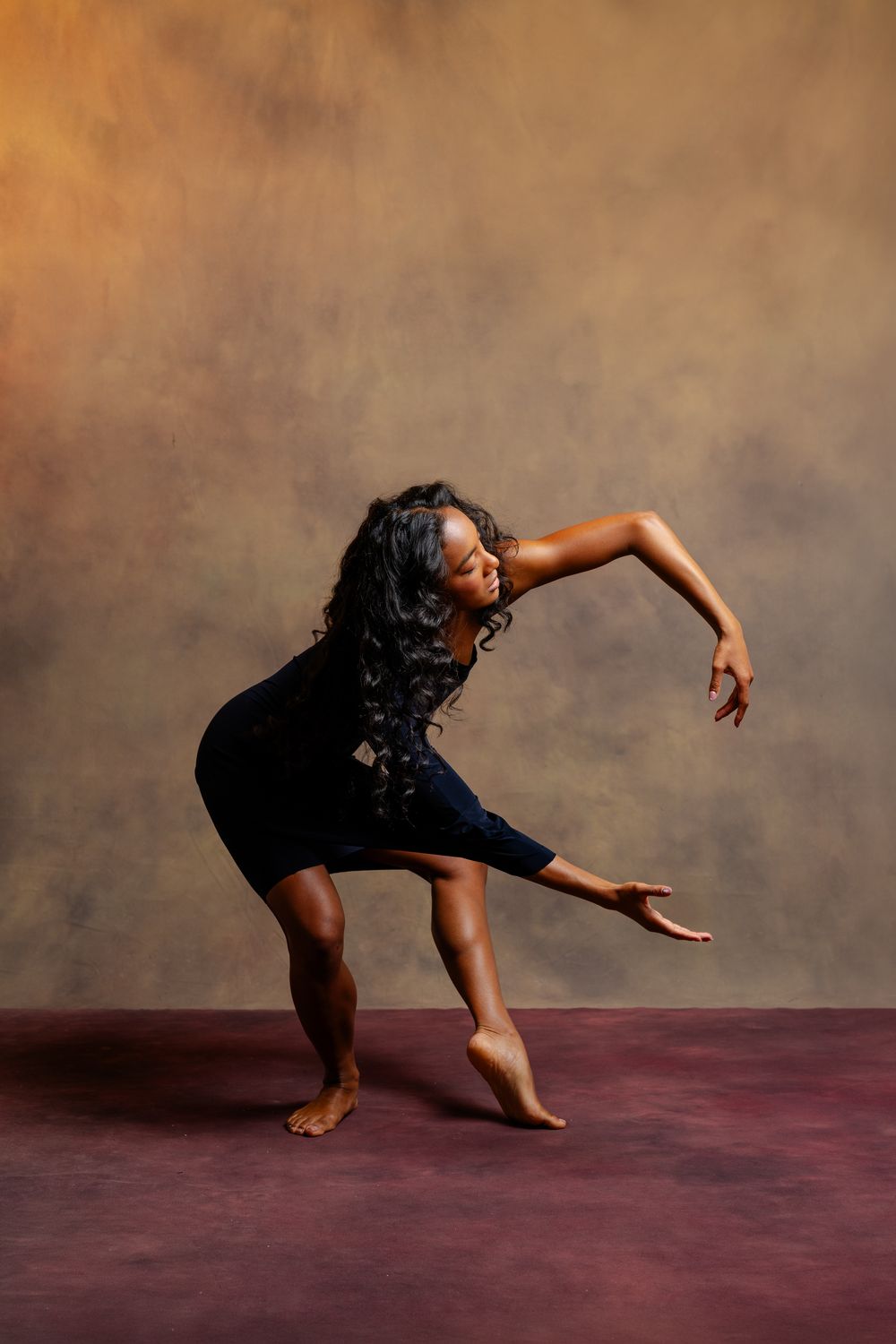 A Black female dancer in a navy dress strikes a dramatic bent pose with flowing hair against a warm studio backdrop.