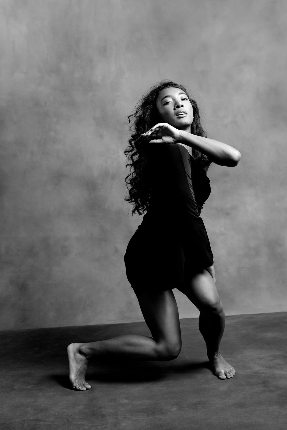 Black and white photo of a young woman in a dance pose, kneeling gracefully with hands clasped, wearing a black outfit.
