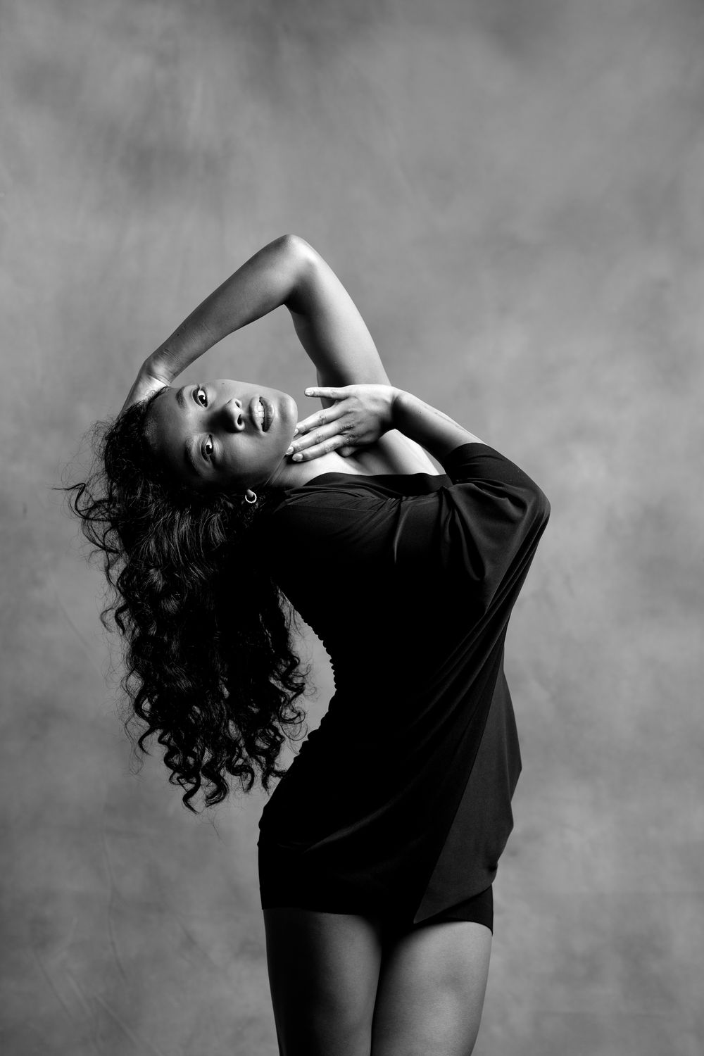 Black and white photo of a woman in a black dress posing gracefully with hands raised and long curly hair flowing.