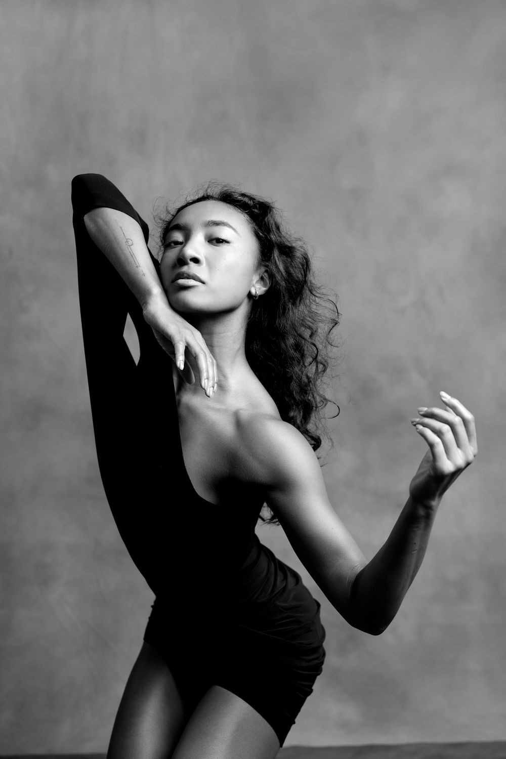 Black and white photo of a flexible female dancer in a leotard performing a high leg extension pose in a studio.