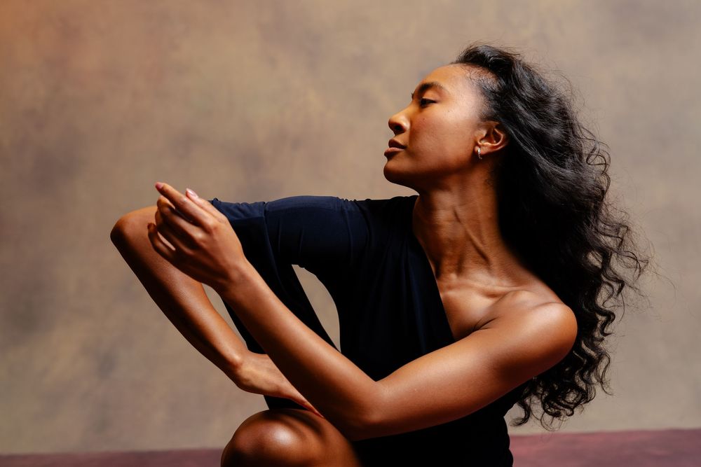 A woman in a black outfit sits cross-legged, eyes closed, hands clasped in a serene meditative pose.