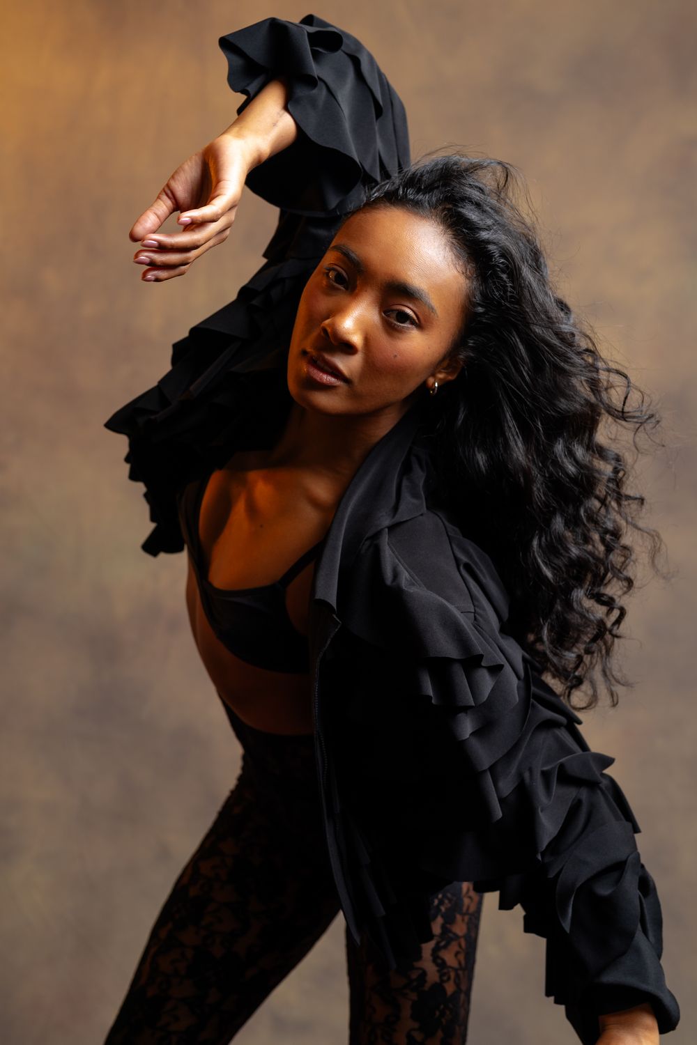 Woman in black ruffled jacket and lace leggings posing dynamically with arm raised in studio.