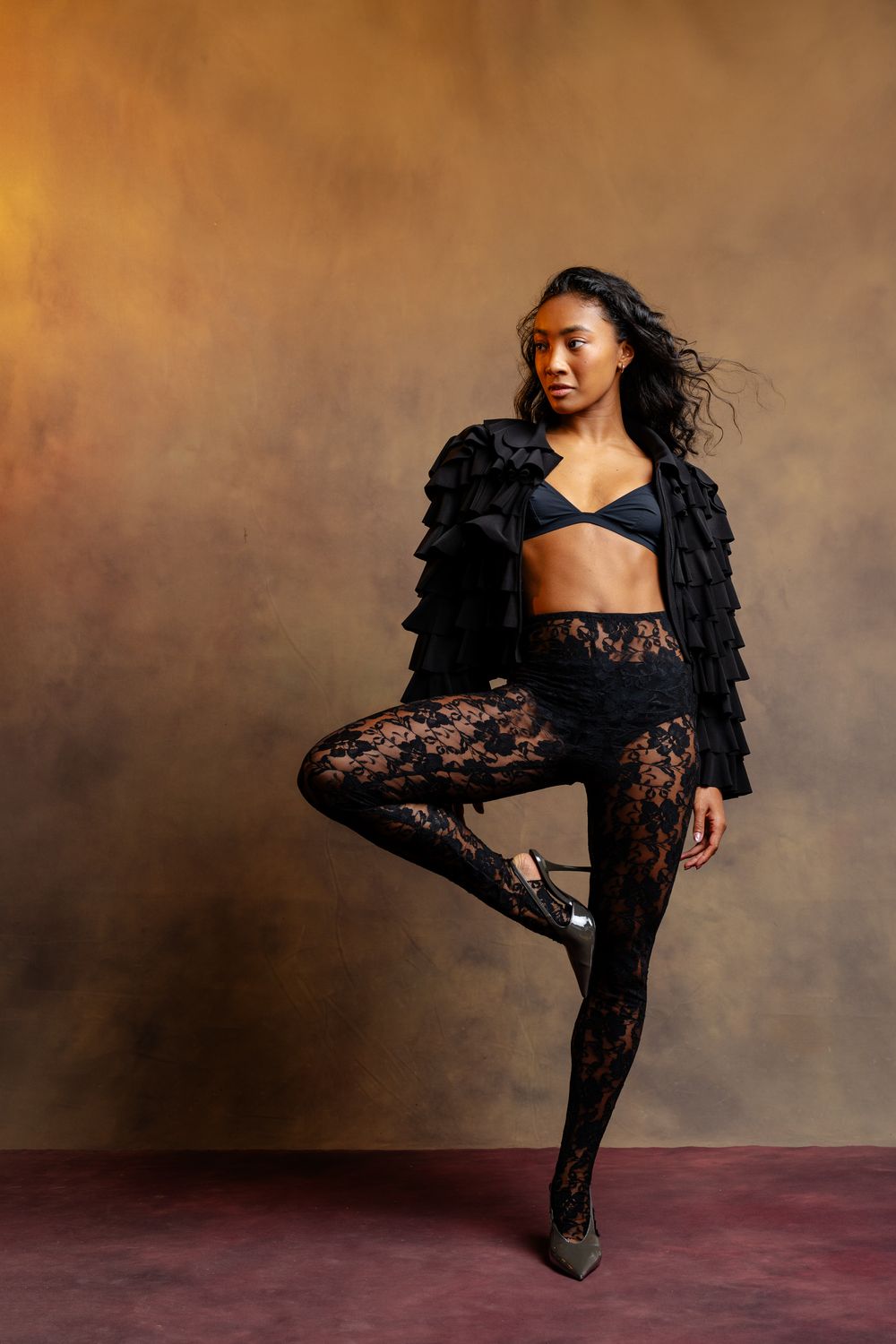 Model in black ruffled jacket and lace leggings balancing on one leg in studio setting.