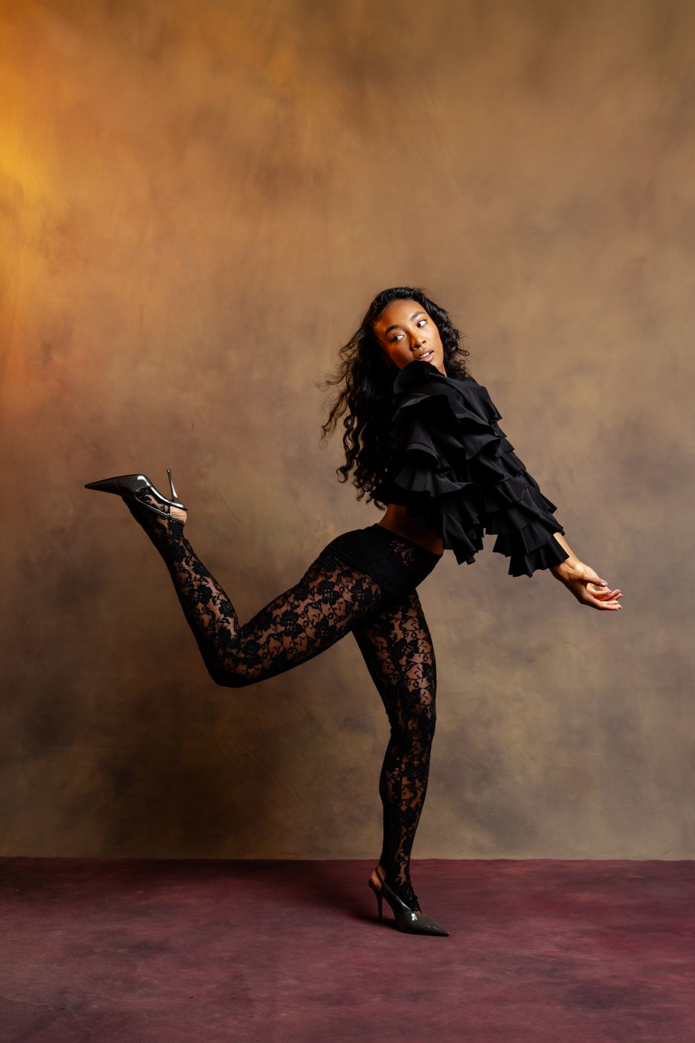 A dancer in black lace tights and ruffled top poses dynamically on one leg in a warmly lit studio.