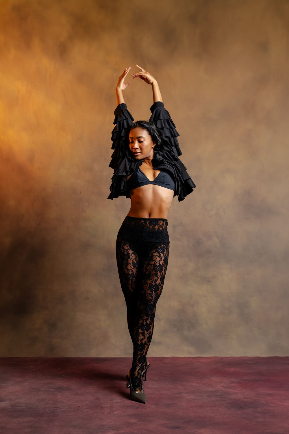 A dancer in black ruffled top and lace pants poses elegantly with arms raised overhead on a golden background.