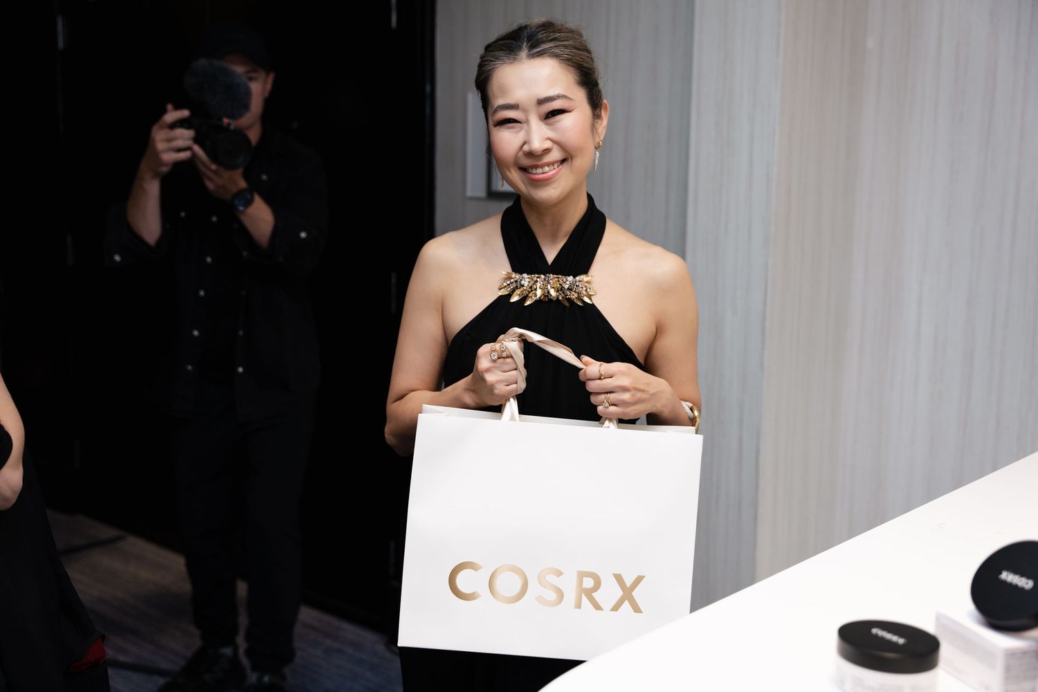 Woman smiling and holding a white COSRX shopping bag at a beauty brand event.