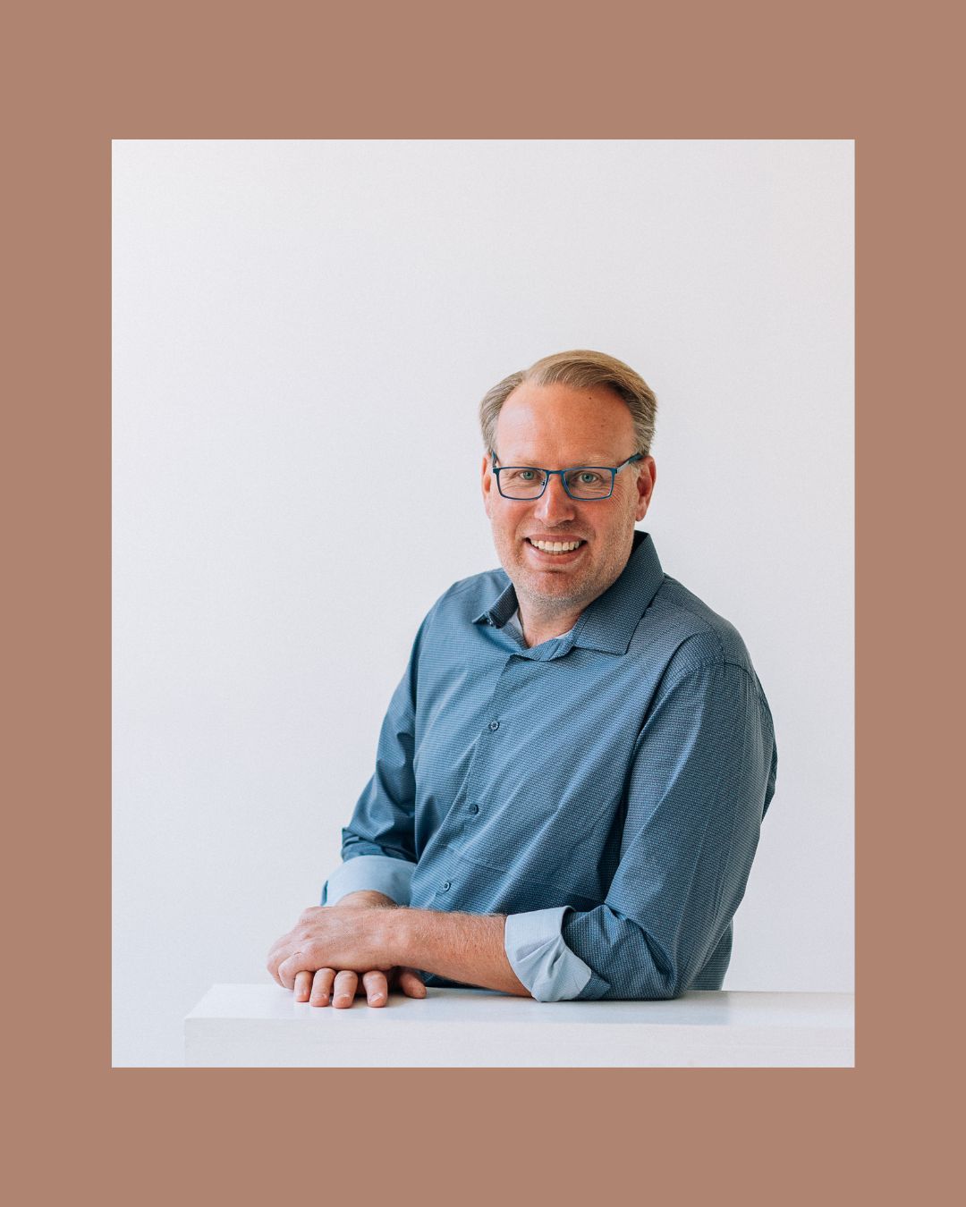 A smiling man with glasses wearing a blue shirt leans on a white surface against a clean white background.