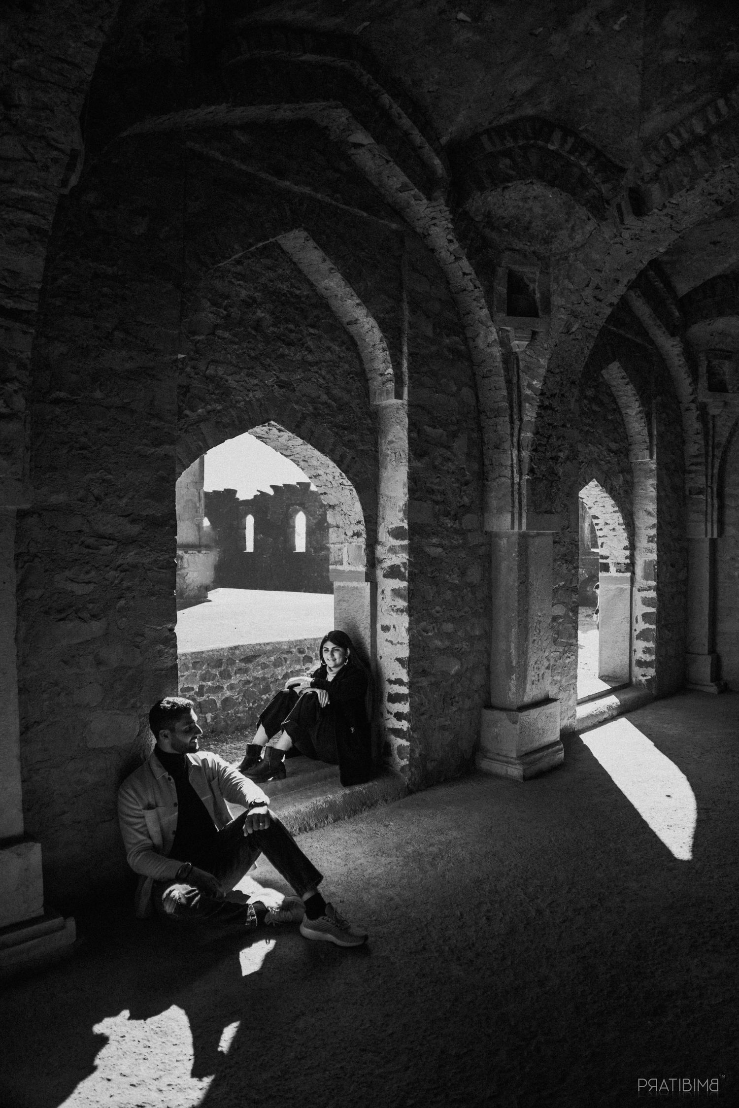 Black and white photo of couple relaxing under ancient stone arches at a historic monument.