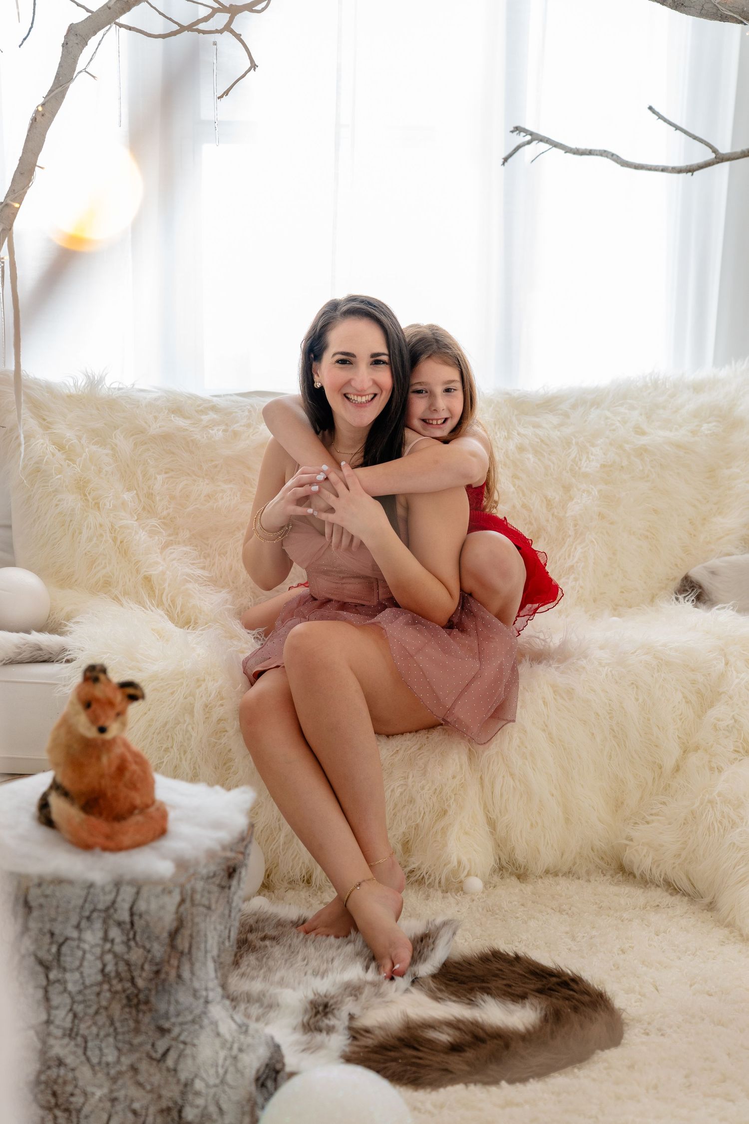 A smiling mother and daughter in dresses share a warm hug on a fluffy white sofa in a bright holiday setting.