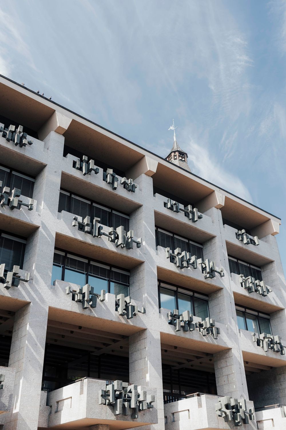 Brutalist concrete building facade with multiple balconies and air conditioning units against a blue sky.