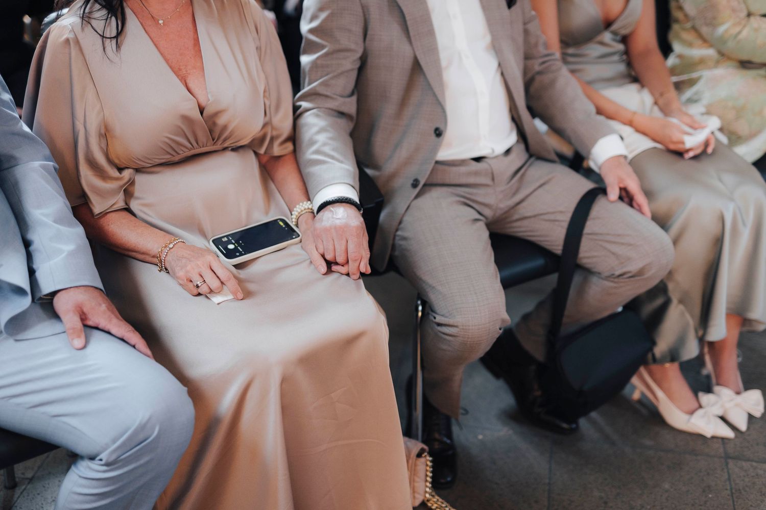 Elegant couple in matching taupe outfits sitting together at a formal event, woman holding a smartphone.